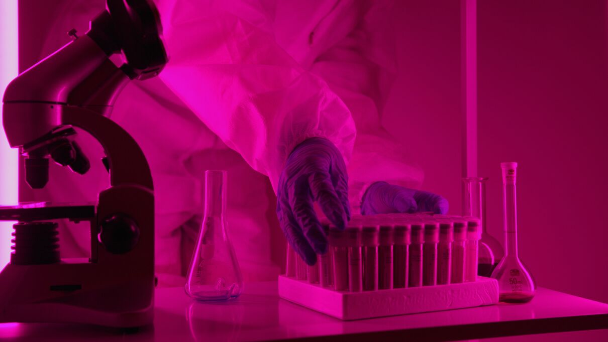 A pink-hued science lab featuring a microscope and gloved hands handling a collection of vials.